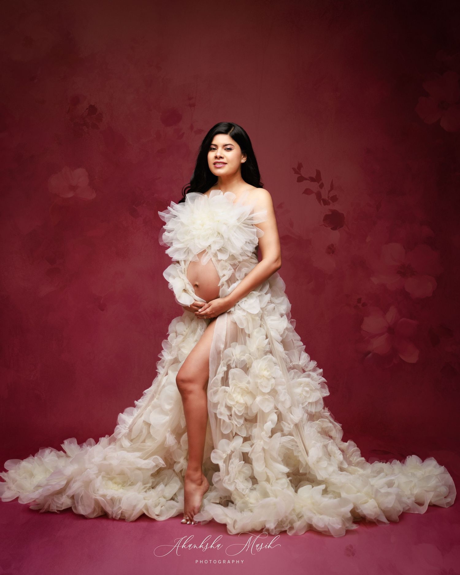 Woman in elegant ruffled white gown poses against deep red studio backdrop.