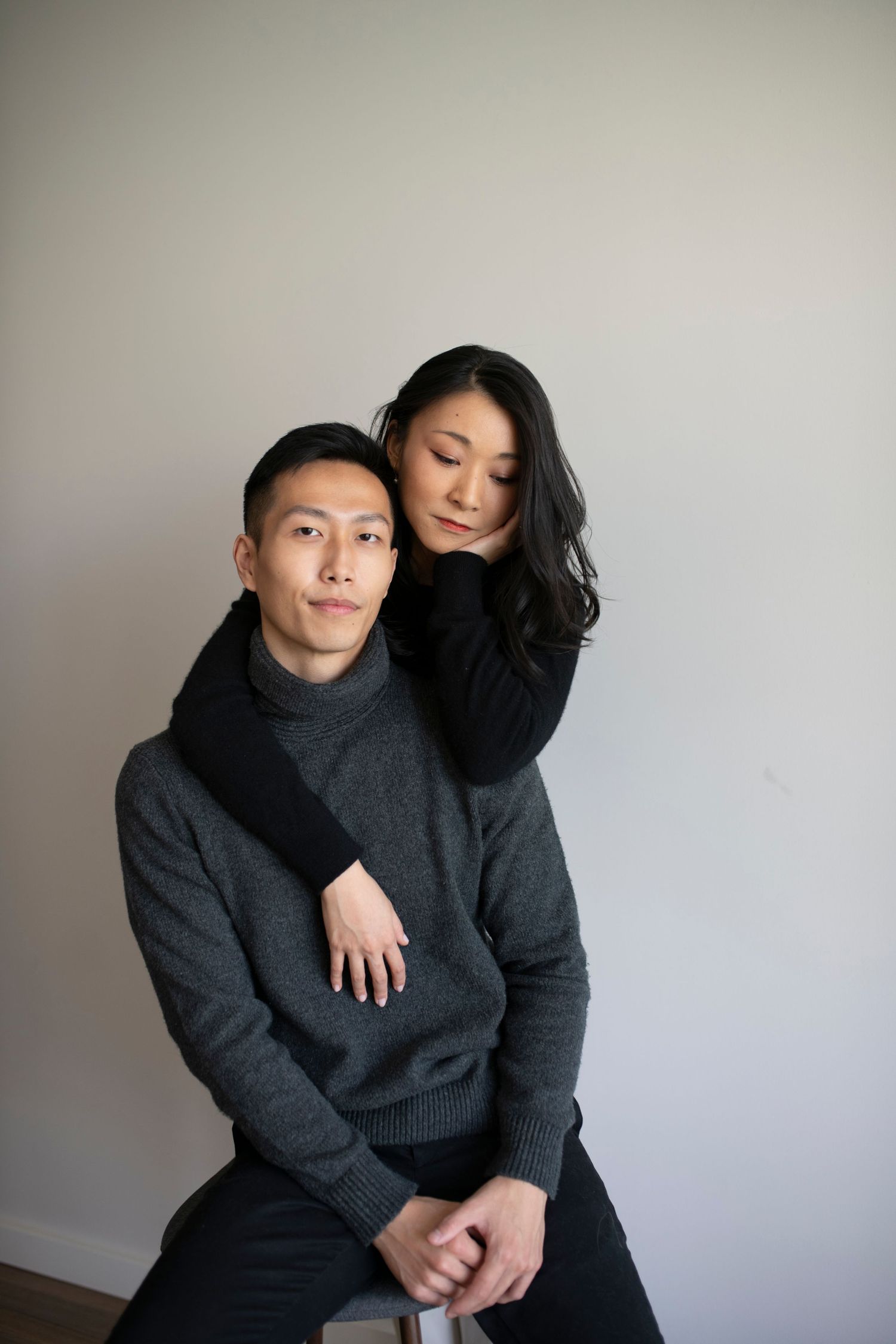 Couple in dark sweaters embracing in a studio portrait, she standing behind him with arms wrapped around his shoulders.
