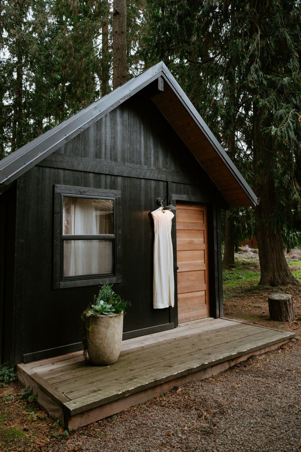 A rustic black cabin with a white dress hanging outside against a backdrop of evergreen trees.