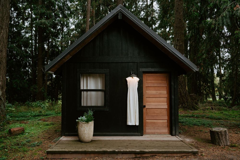 A minimalist black wooden cabin with a white dress displayed on its front porch.