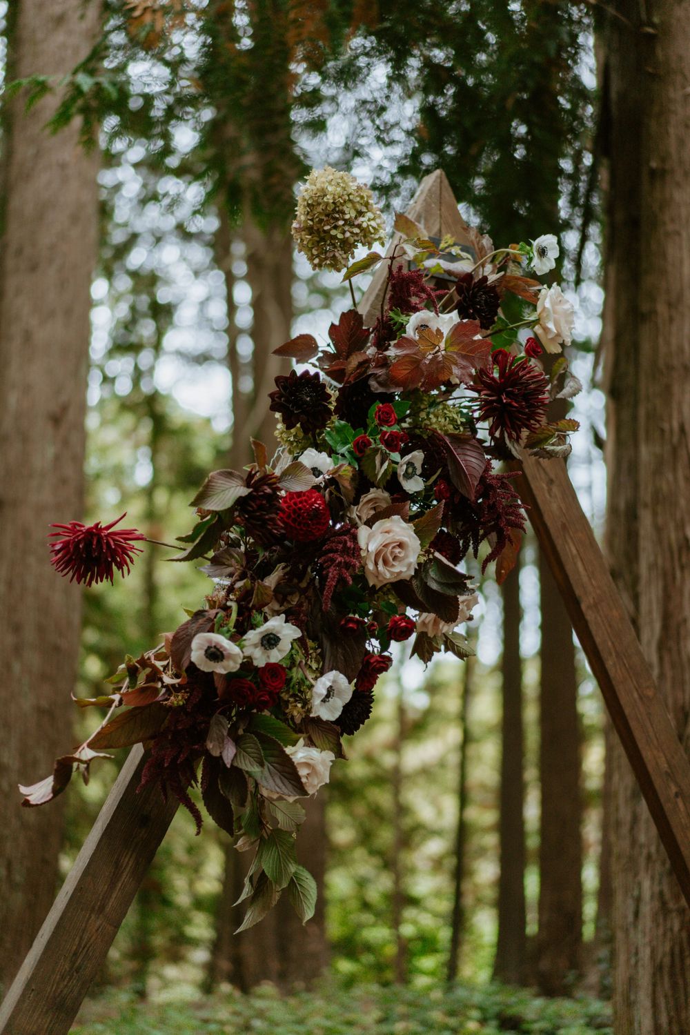 Rustic wooden arch adorned with deep burgundy and blush floral arrangements in a forest setting.