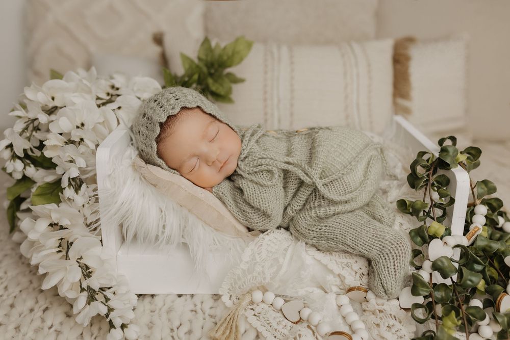Peaceful newborn sleeping on white bedding with soft green knit wrap and floral accents.