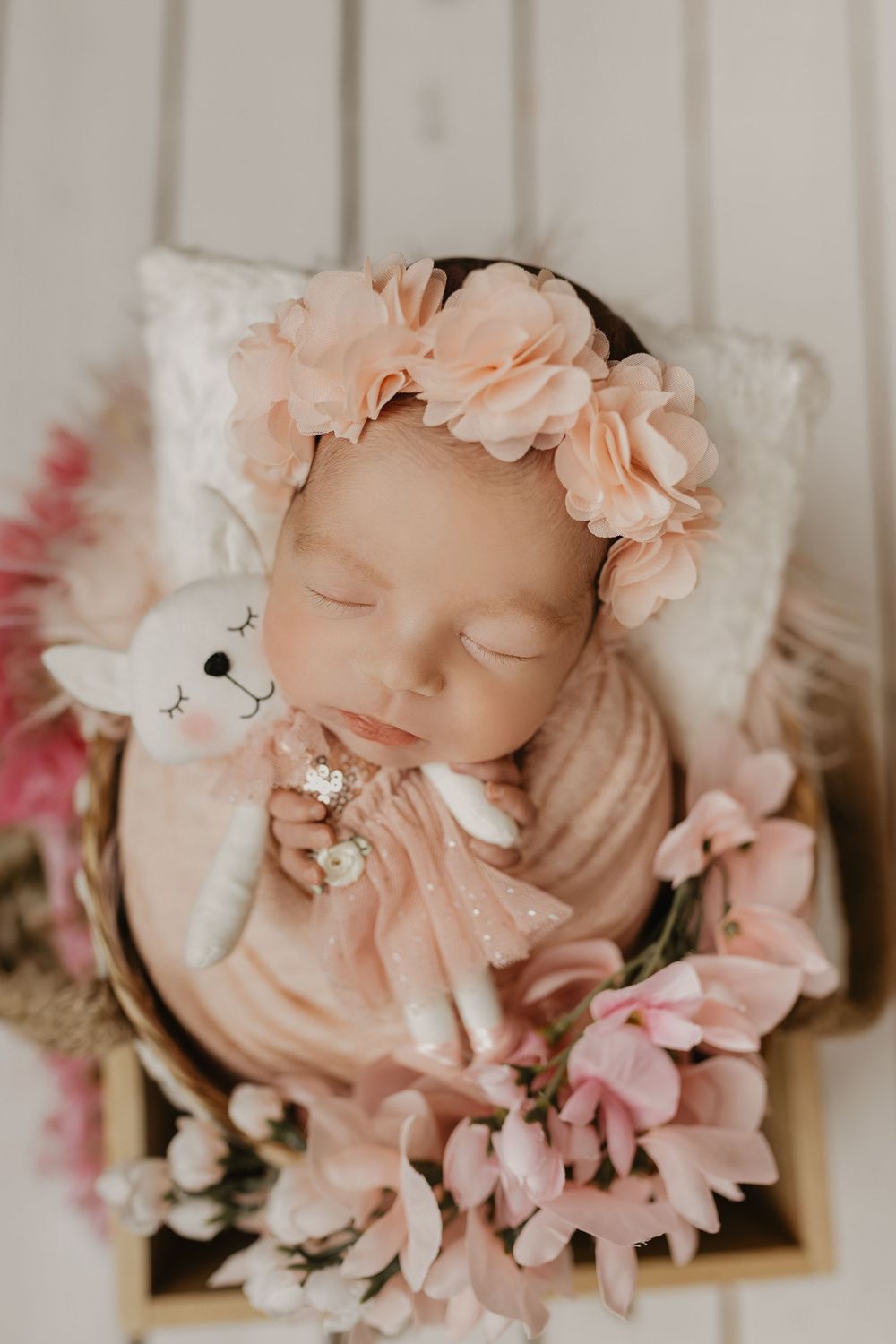Dreaming newborn adorned with pink flower crown and delicate pearls on white backdrop.