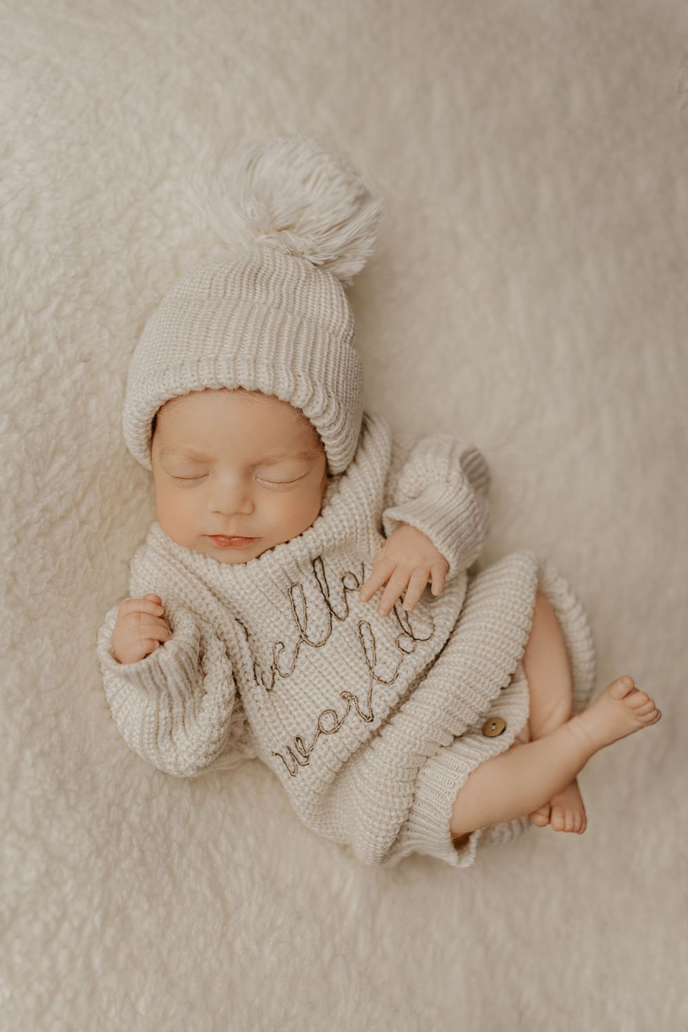 Sleeping baby in cream knitted outfit on plush white textured background.
