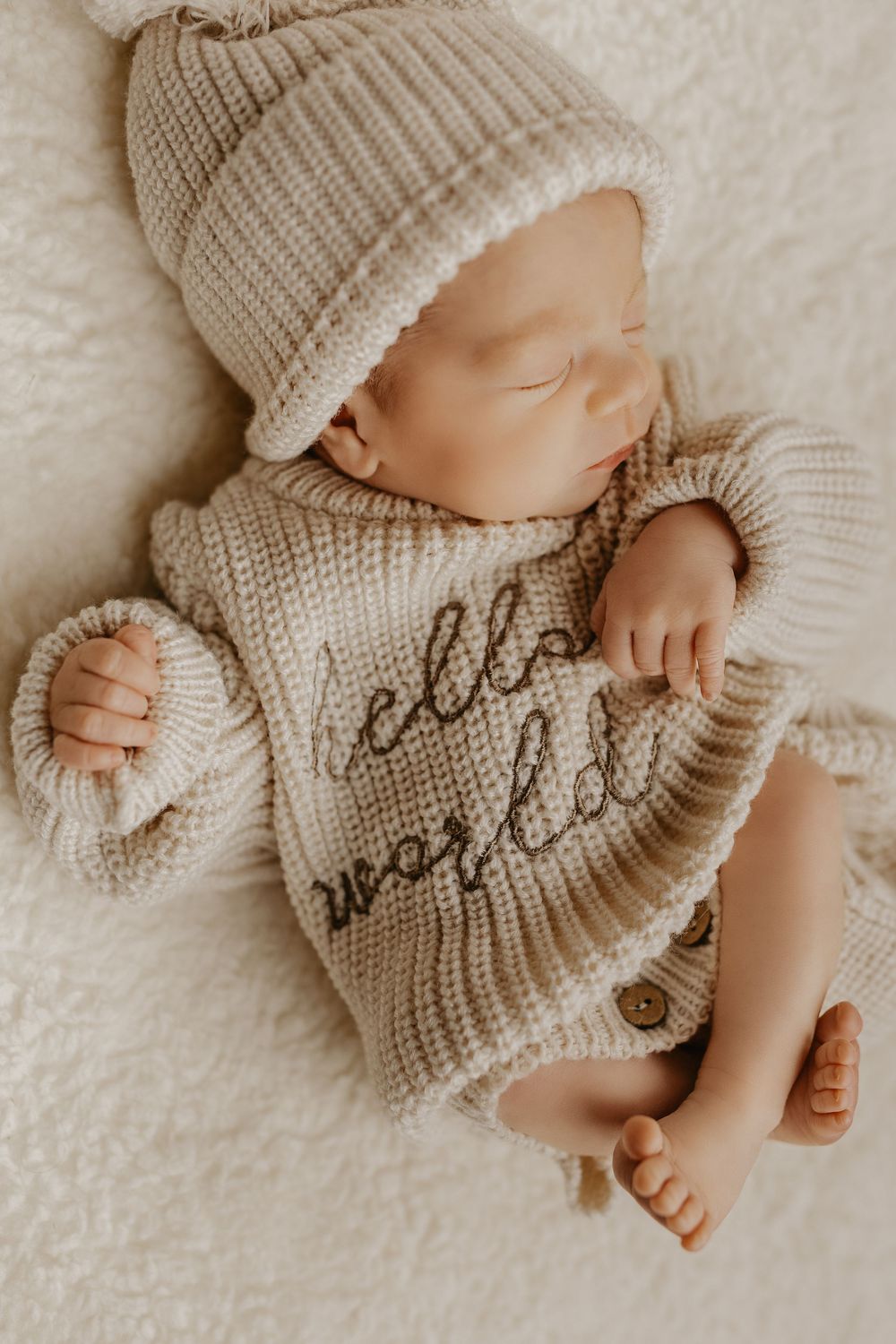Newborn in beige knit sweater and matching hat resting peacefully on white surface.