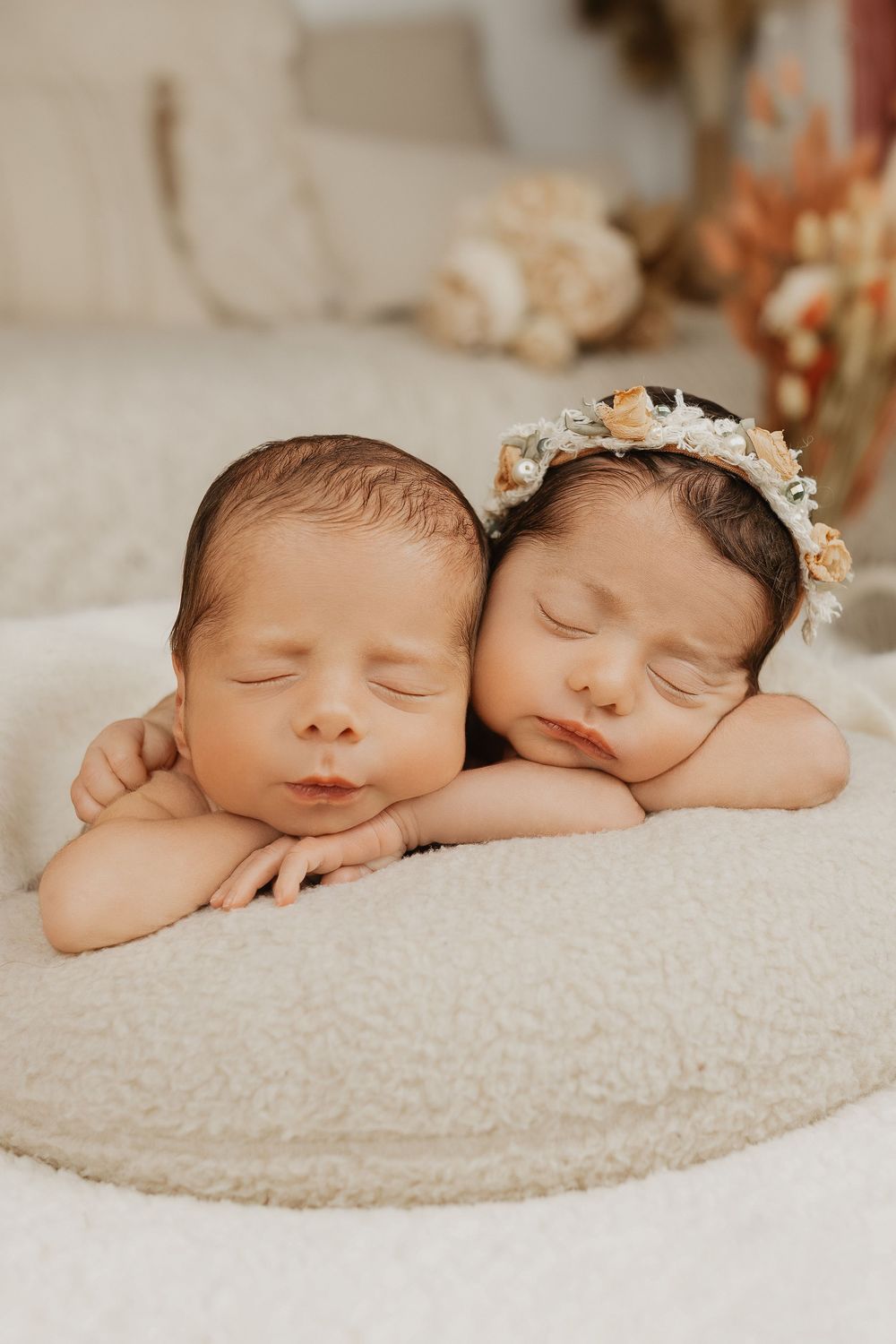 Two sleeping newborn twins rest cheek-to-cheek on a soft white cushion, one wearing a delicate floral headband.