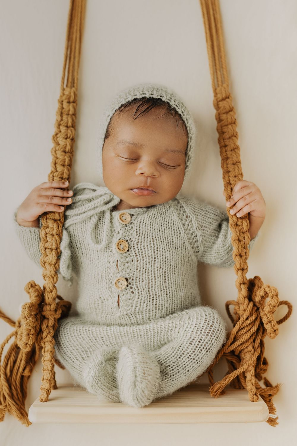 Sleeping newborn baby in sage green knit outfit on macrame swing.