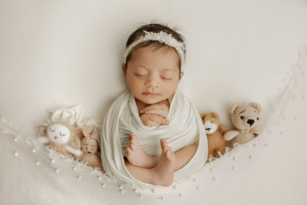 Sleeping newborn with lace headband surrounded by small plush toys on white backdrop.