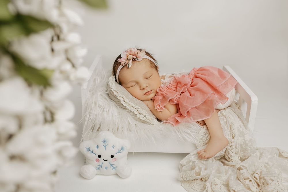 Sleeping newborn in pink outfit surrounded by white lace and floral decorations.