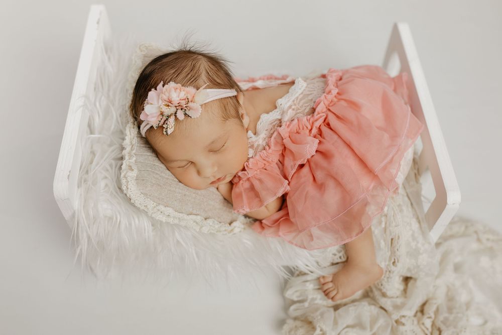 Baby wearing coral pink dress resting on vintage white lace bedding.