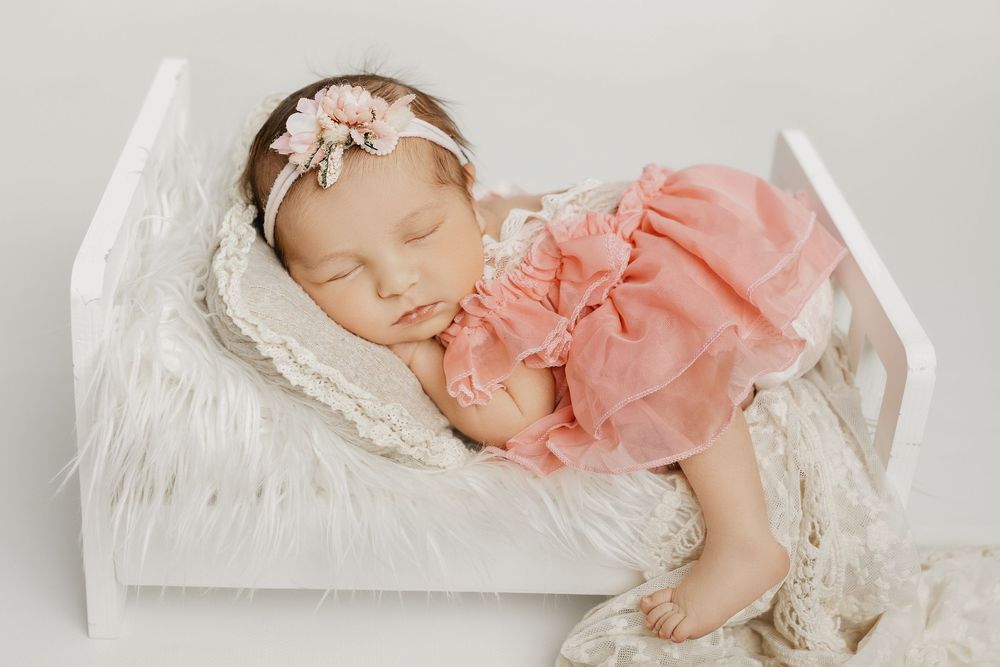 Peaceful newborn in salmon pink dress sleeping on white lace backdrop.