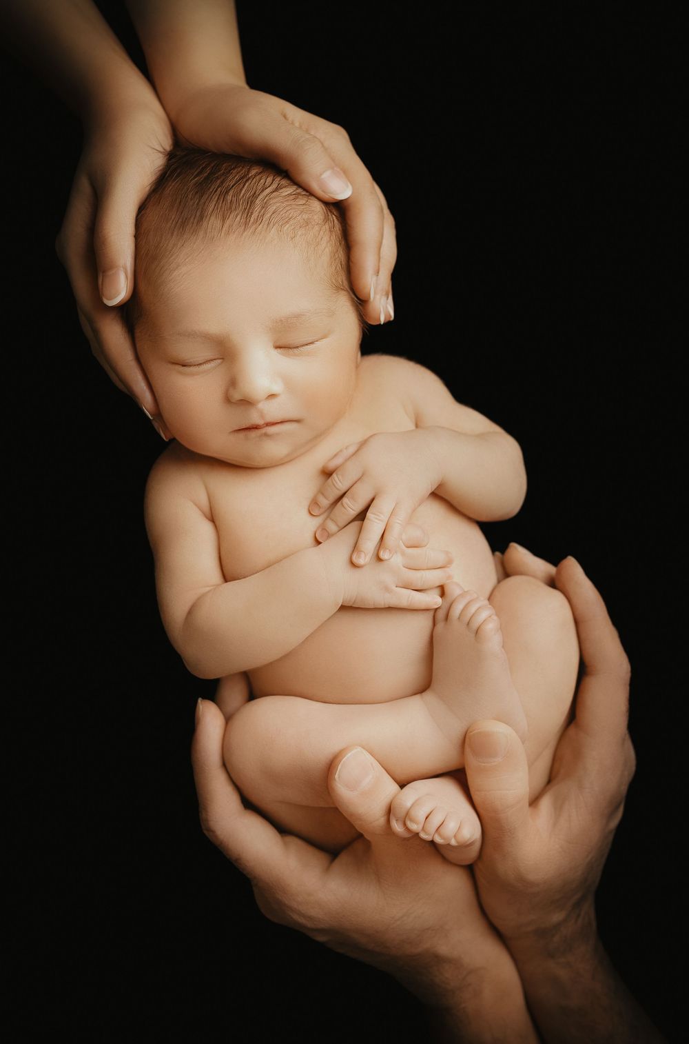 Gentle hands cradling sleeping newborn against dark background in artistic portrait.
