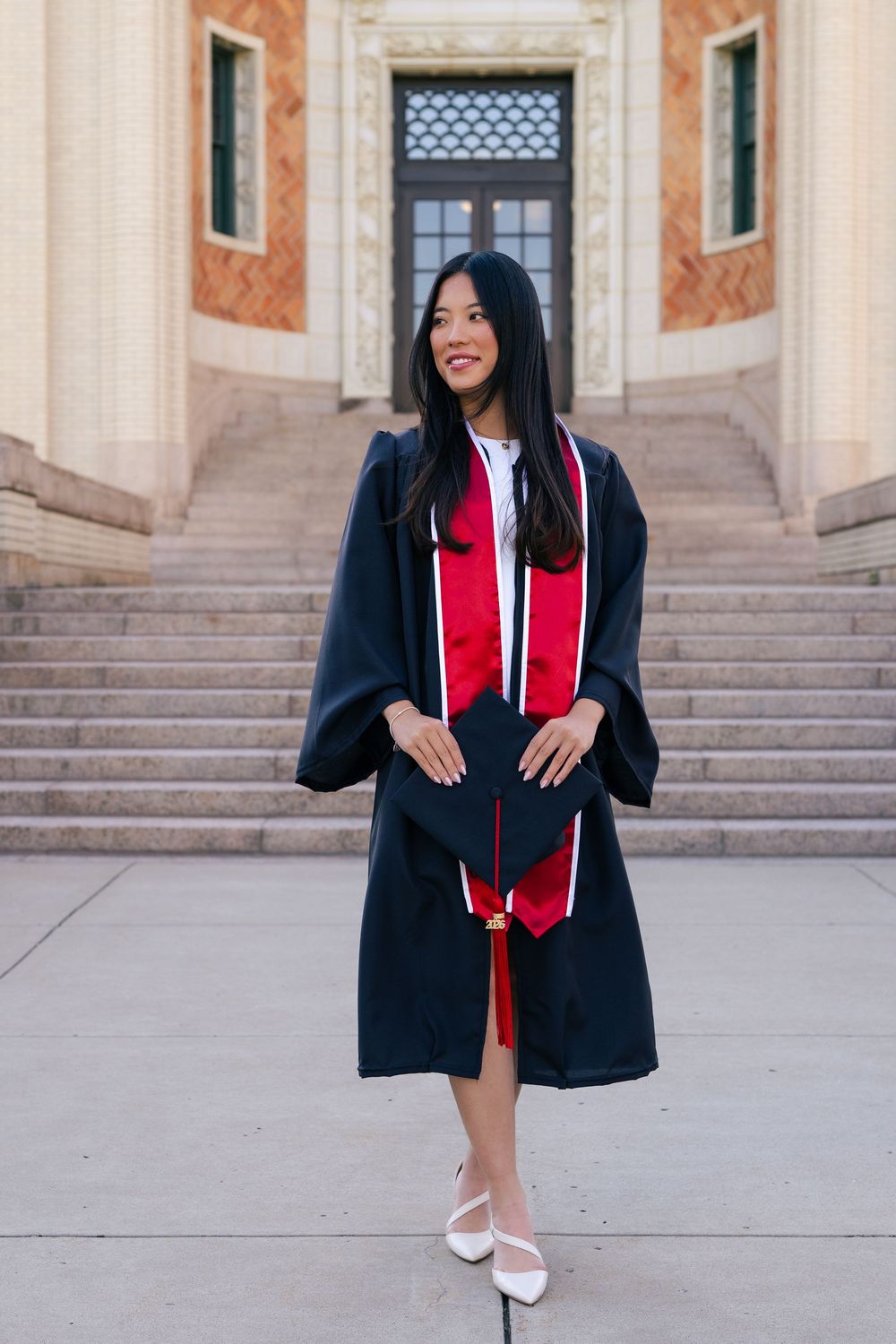 Young woman in black graduation gown with red stole smiles proudly on university steps.