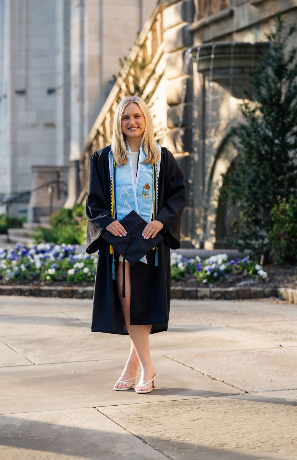 Smiling graduate in black cap and gown with light blue stole poses outdoors near stone building and flower garden.
