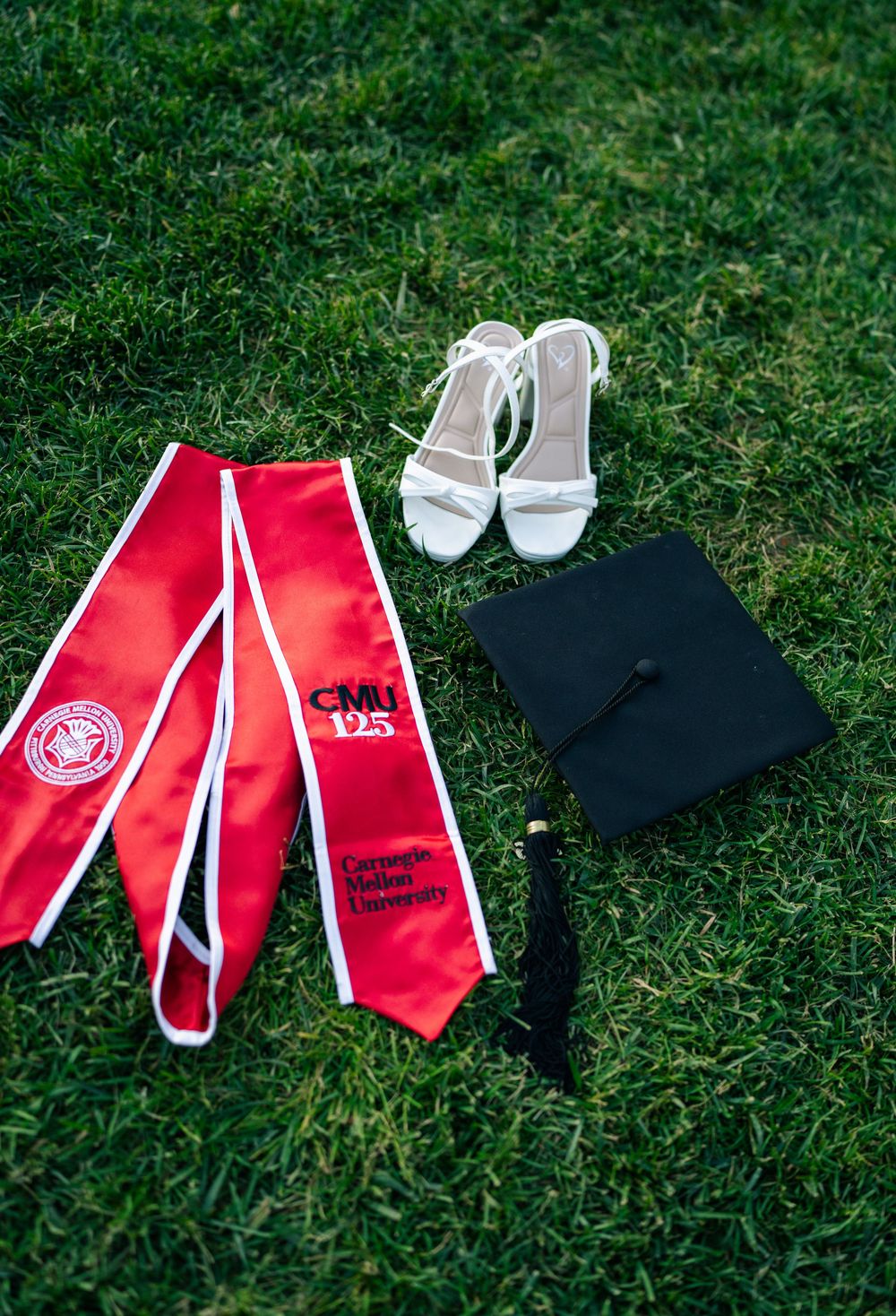 Carnegie Mellon University graduation cap, red CMU 125th anniversary stole, and white heeled sandals arranged on green grass.
