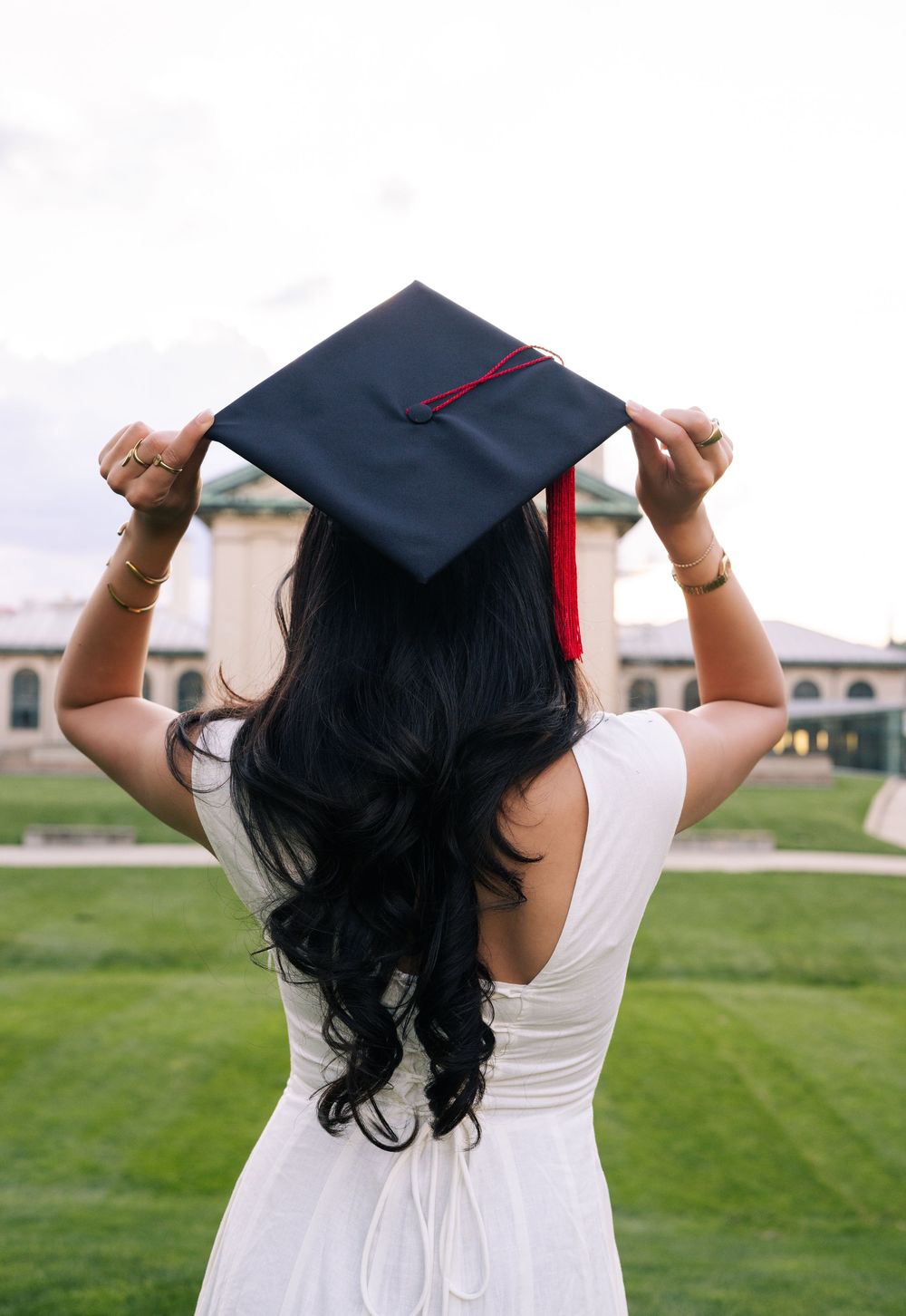 A woman in a white dress holds a black graduation cap with red tassel on her head, viewed from behind on a green campus.