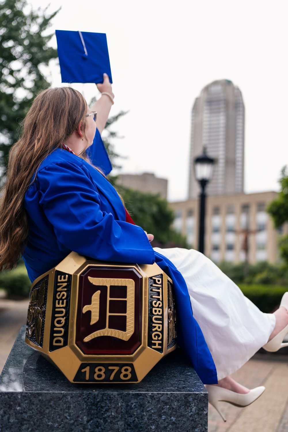 Duquesne University graduate in blue gown holding cap aloft, seated on school emblem monument, Pittsburgh 1878.