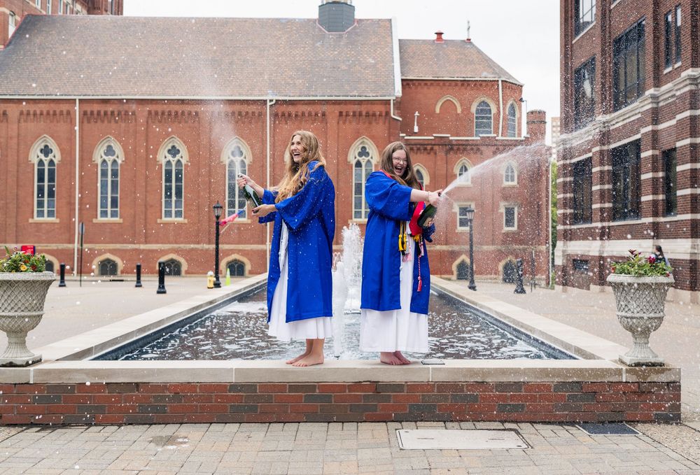 Two graduates in blue robes celebrate with champagne by a fountain outside a red brick university building.
