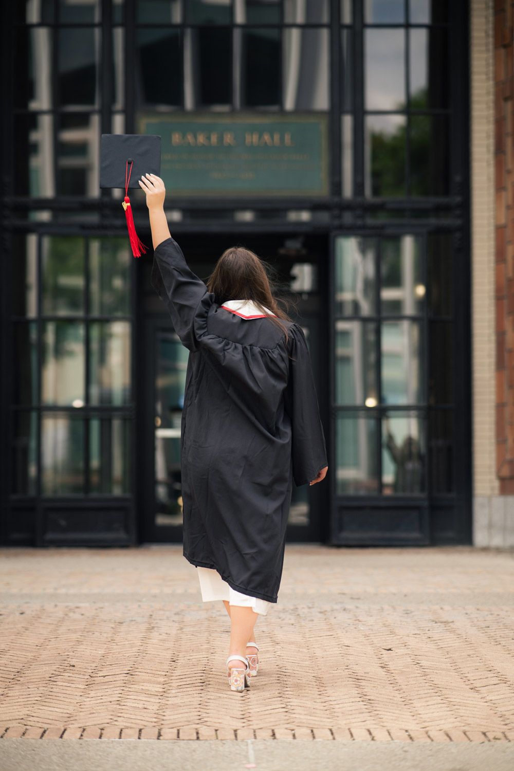A graduate in cap and gown raises their diploma victoriously in front of Baker Hall on a brick walkway.