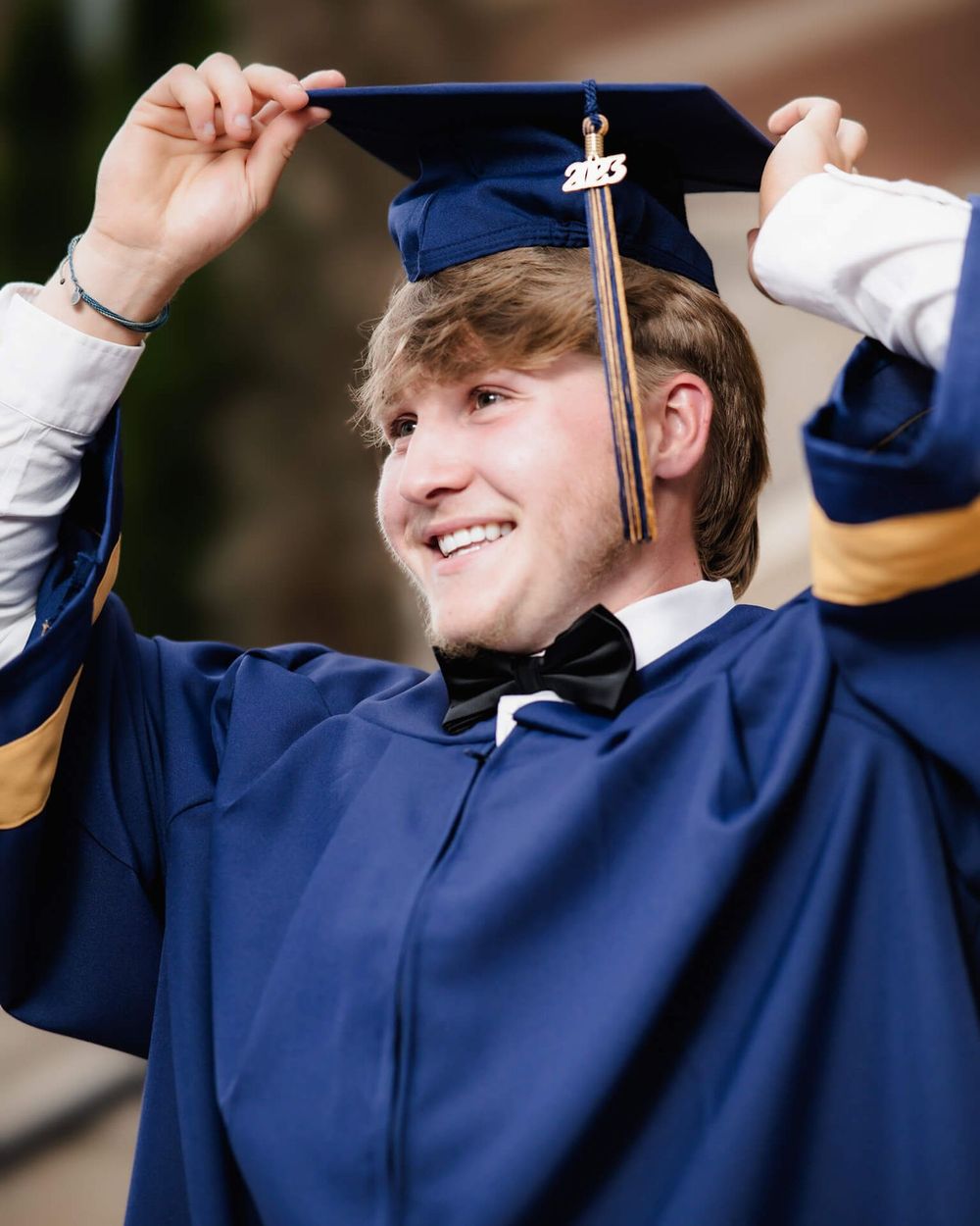 Graduate in navy blue cap and gown adjusts mortarboard tassel during graduation ceremony while smiling.