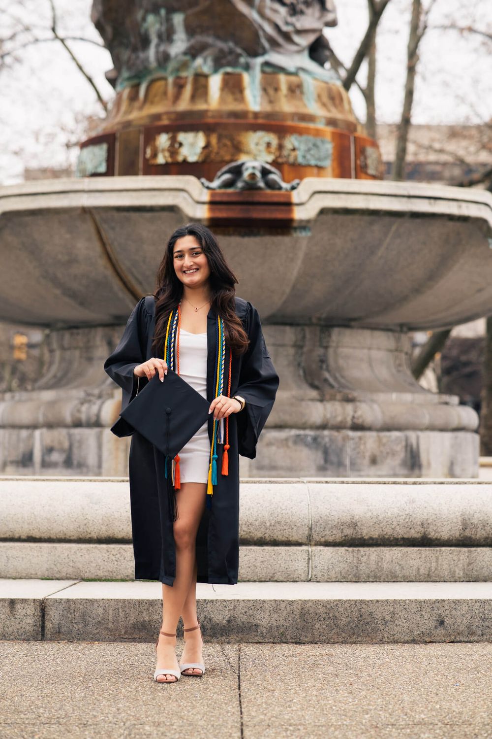 Graduate in cap and gown poses in front of ornate stone fountain wearing graduation regalia and honors cords.