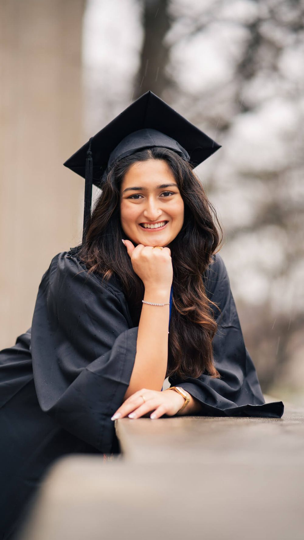 Graduate in black cap and gown poses for outdoor graduation photo with hand resting on chin.