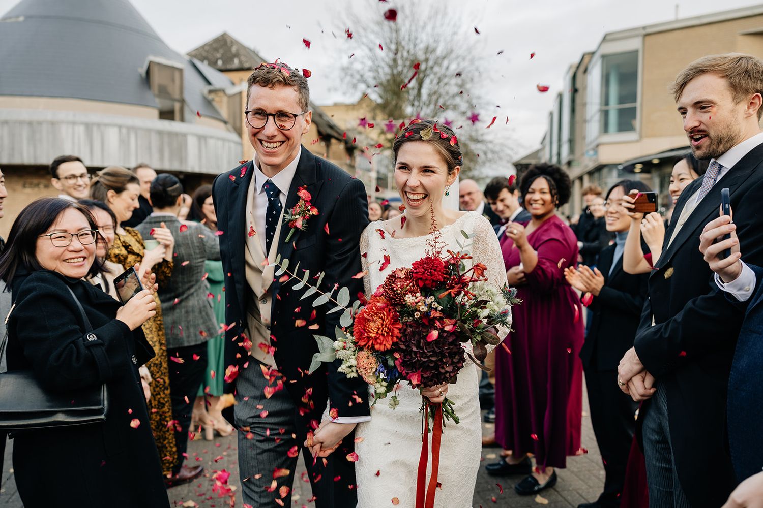 Wedding celebration with guests throwing rose petals as couple walks down aisle holding vibrant red bouquet.