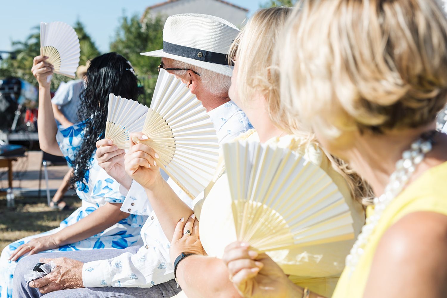 People use white handheld fans to cool off while sitting outdoors on a sunny summer day.