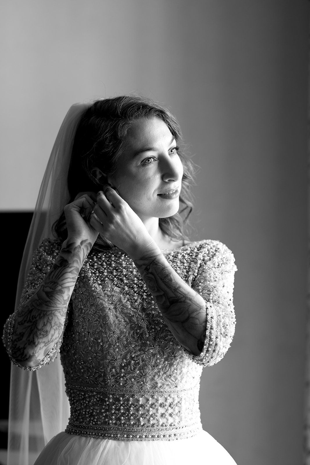 Black and white portrait of a bride in beaded lace wedding dress putting on earrings near a window.