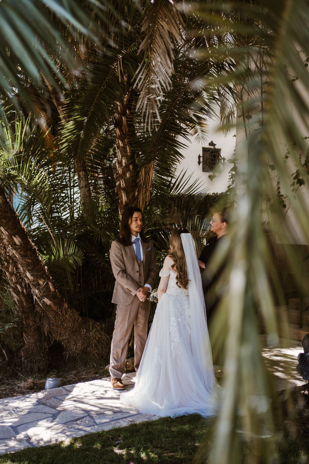 A wedding ceremony moment framed by tropical palm fronds in natural sunlight.