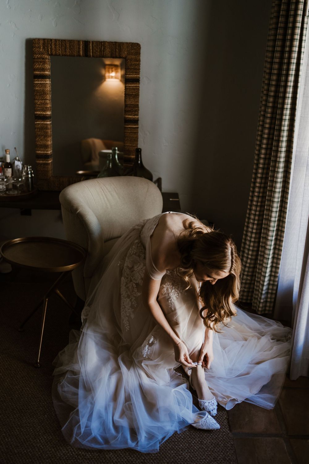 A quiet moment as someone in a white wedding gown adjusts their shoe in a dimly lit room.