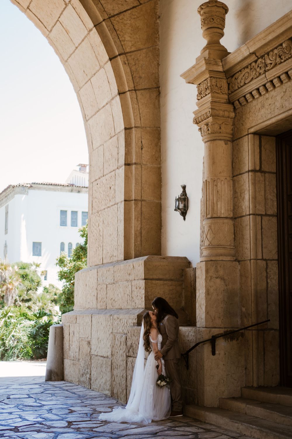 Two figures in wedding attire share an intimate moment under stone archways at an elegant Mediterranean-style venue.