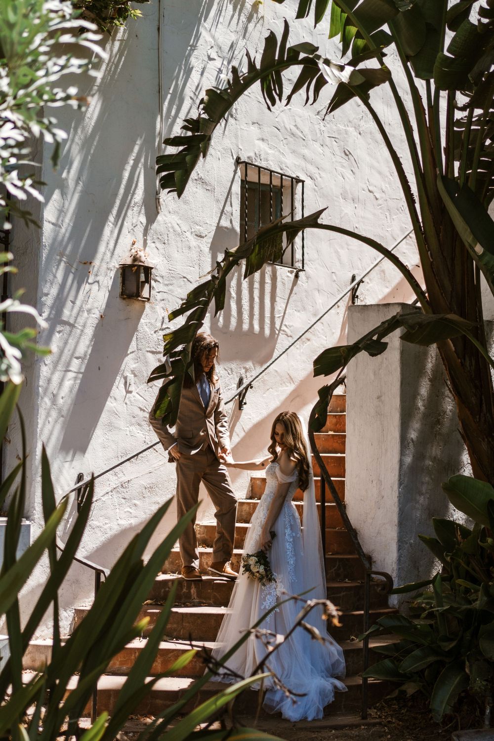Two people stand on white stucco staircase framed by tropical palm fronds in dappled sunlight.