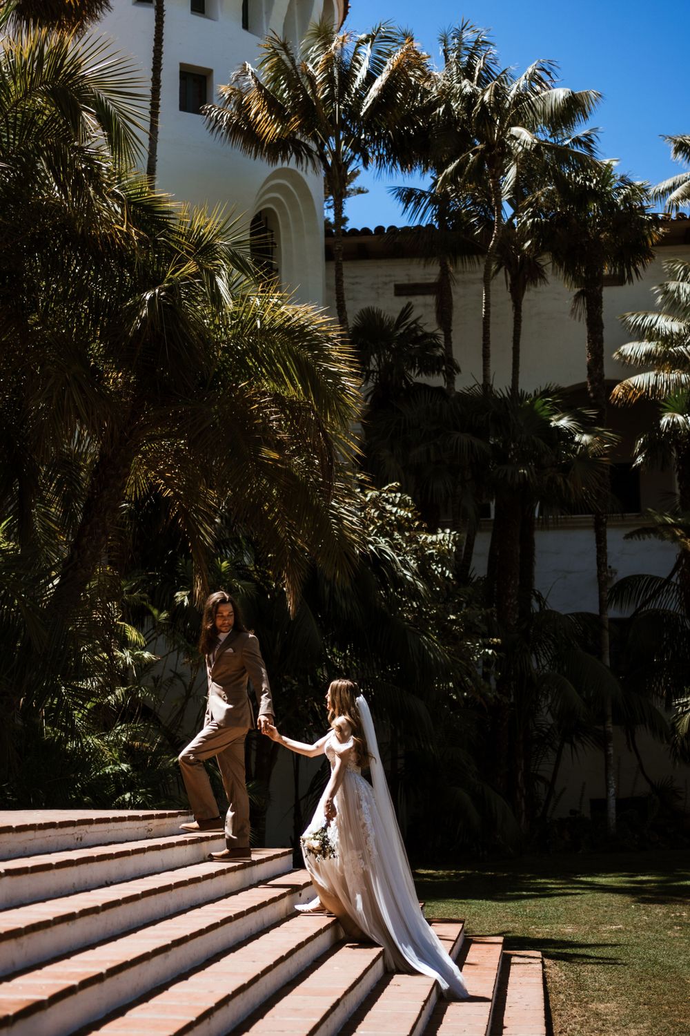 A couple descending grand stone steps at a tropical venue with palm trees and Spanish colonial architecture.
