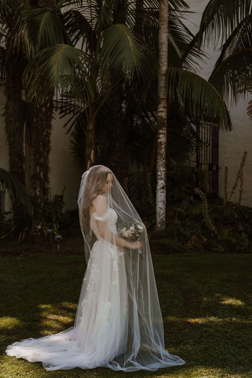 A bride in a flowing white wedding gown and long veil stands elegantly among palm trees in a tropical garden setting.