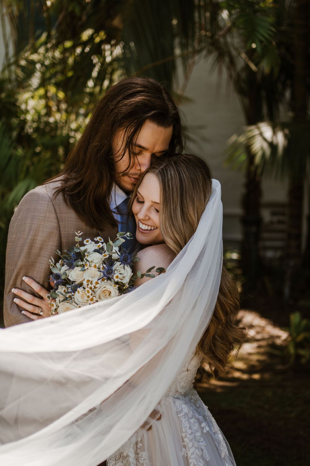 Intimate moment between newlyweds embracing under garden foliage with white veil and delicate floral bouquet.