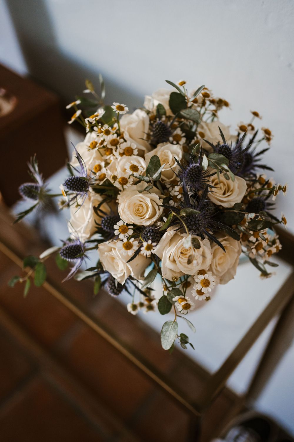 An elegant wedding bouquet with cream roses, blue thistles, and delicate greenery sits on a wooden surface.