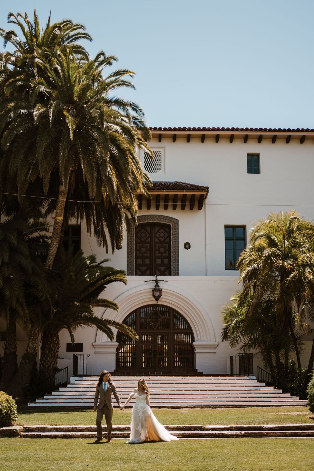 A Spanish Colonial Revival style building with white walls, arched entryway and tall palm trees framing the entrance steps.