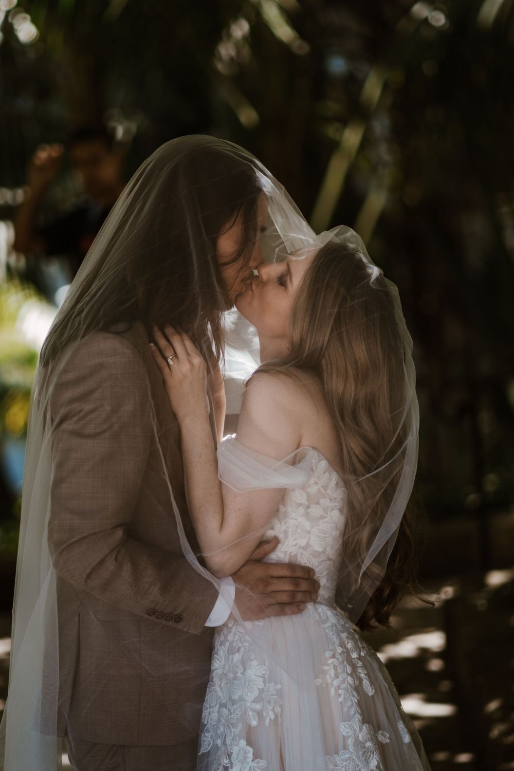 A couple in wedding attire share a romantic kiss under a veil in a sunlit outdoor garden setting.