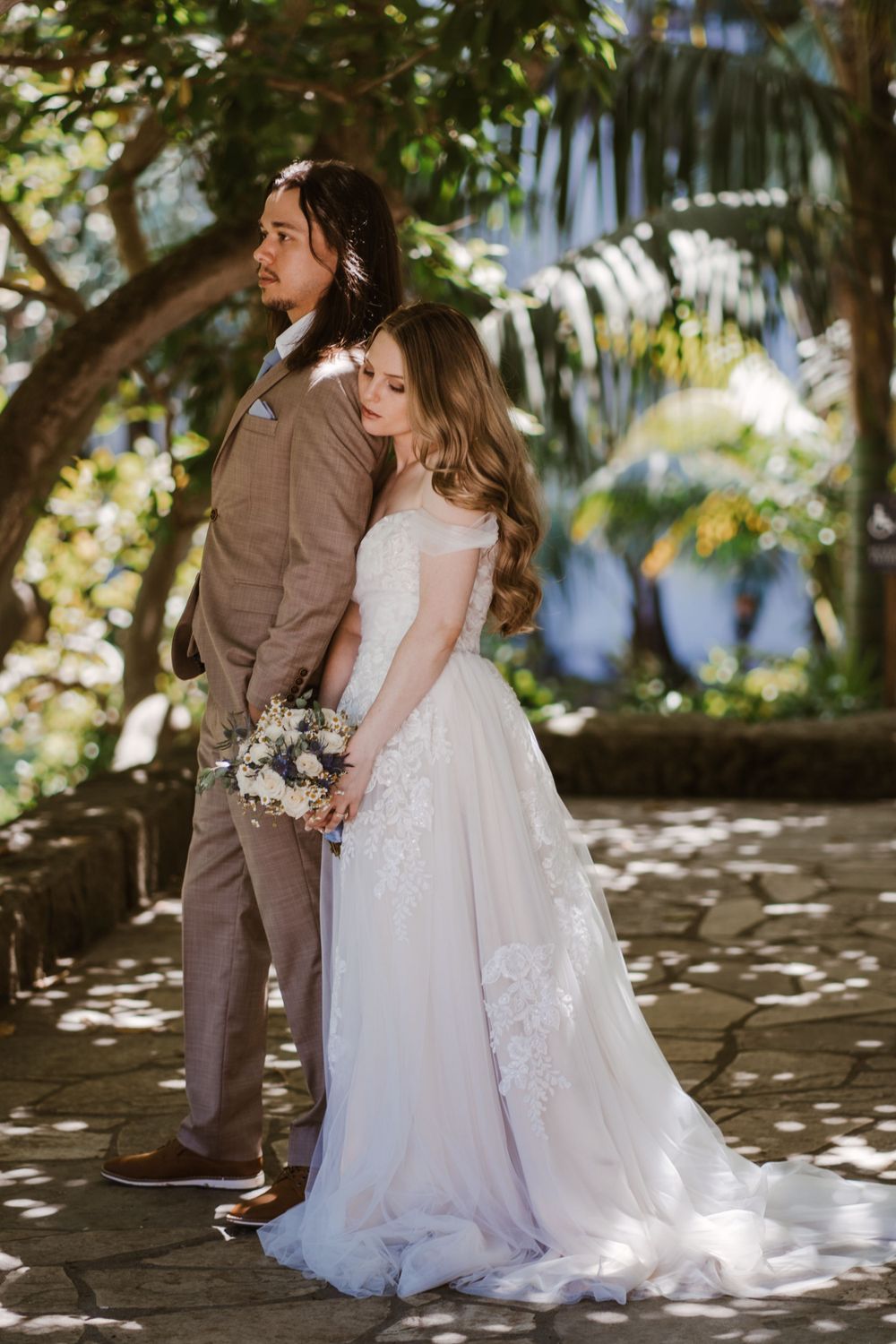 Bride in flowing white dress holds bouquet while standing with groom in tan suit under dappled sunlight in tropical garden.