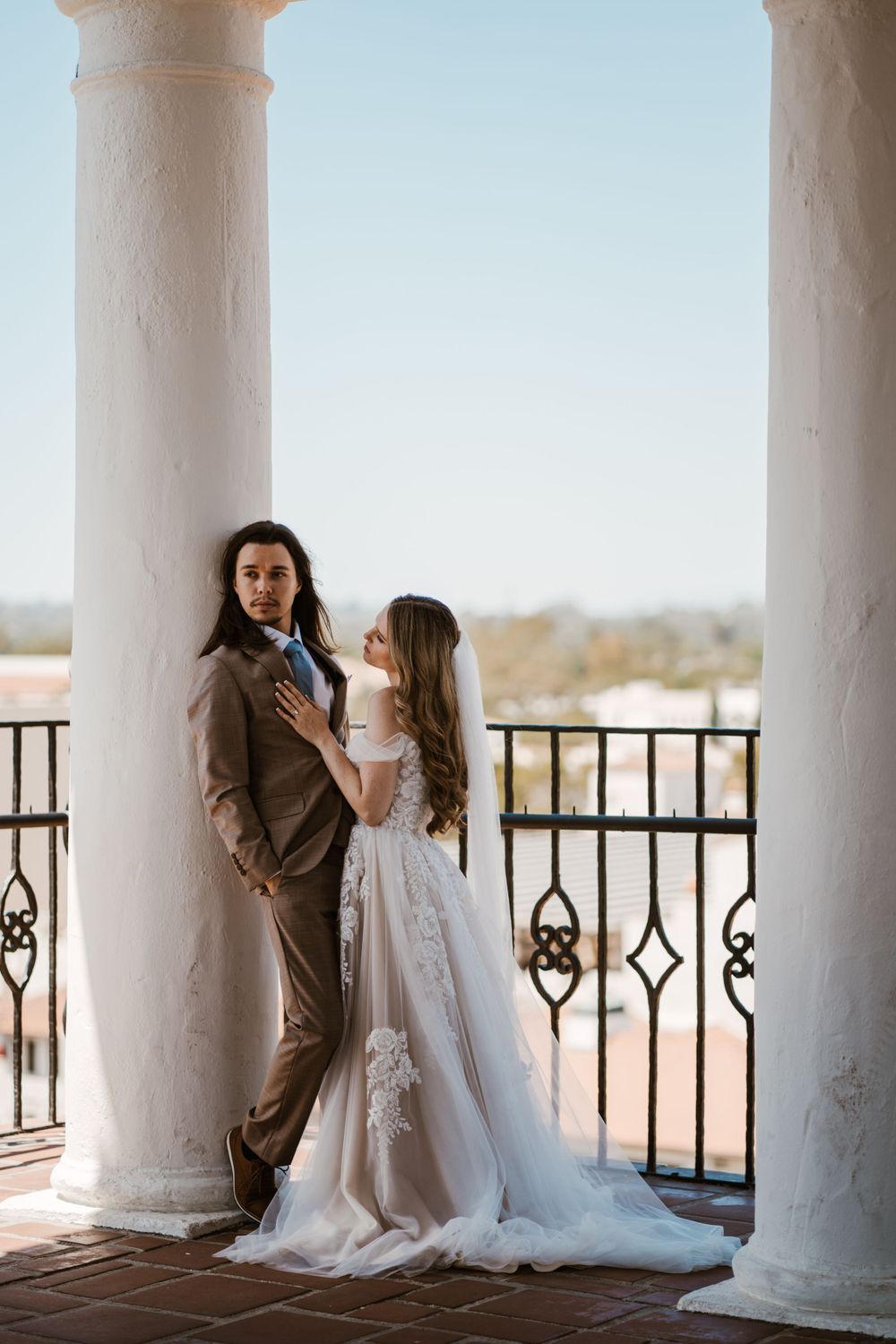 Elegant couple in wedding attire pose between white columns on a balcony with ornate railings at sunset.