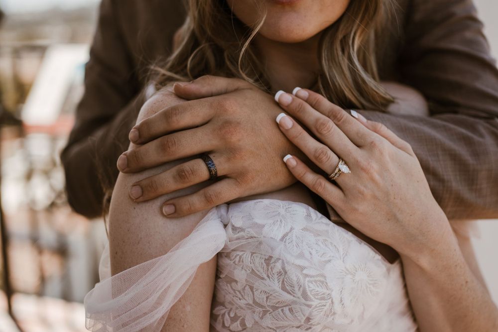 Two hands with engagement or wedding rings shown against white lace dress fabric in romantic portrait.