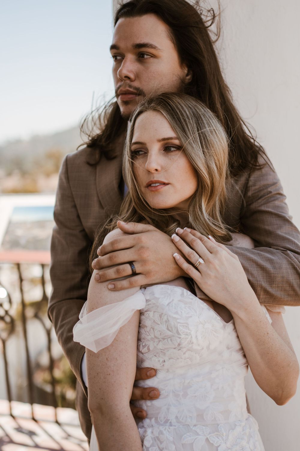 A romantic couple in wedding attire embrace on a balcony, with the person in brown hugging from behind.