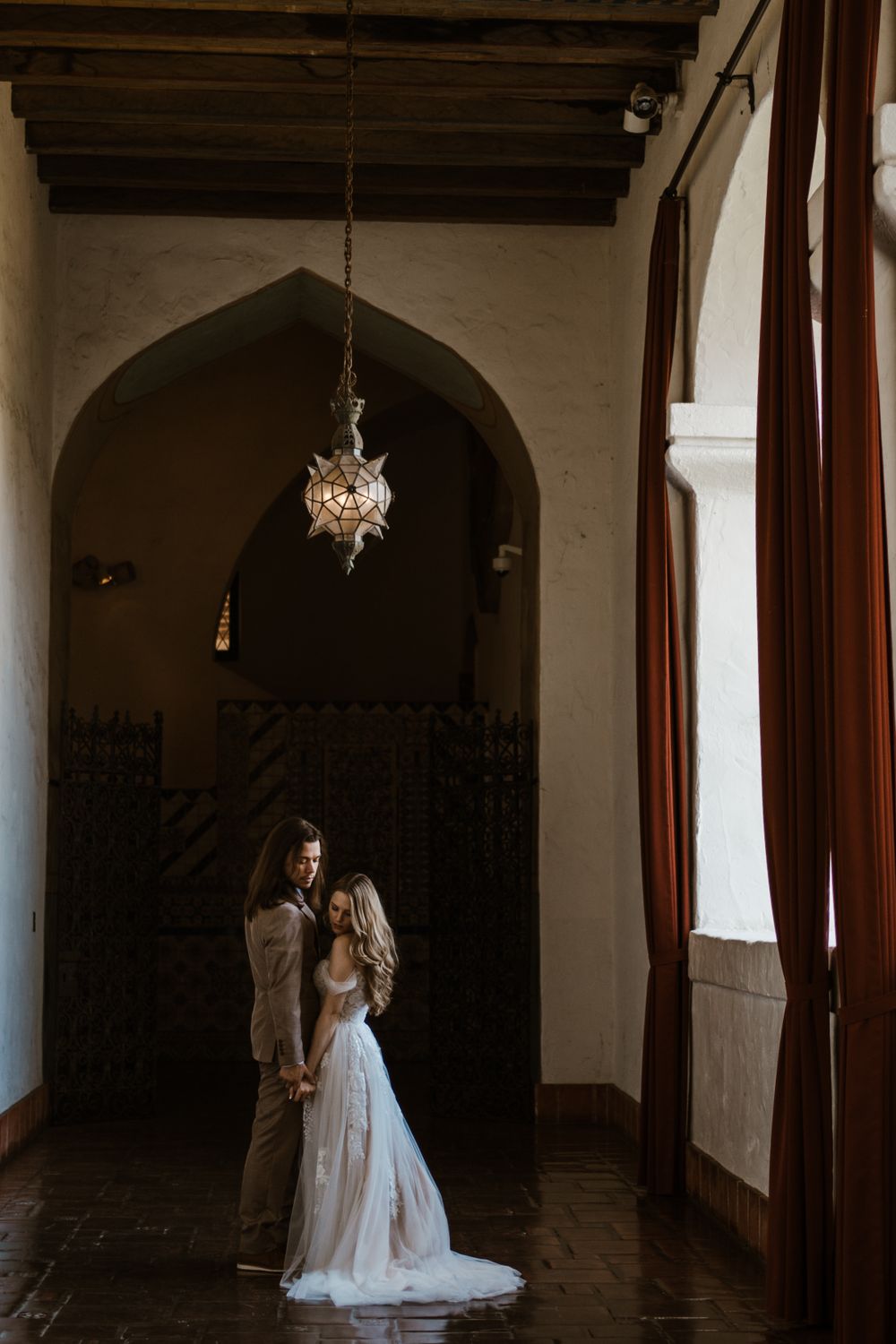 A couple stands in a moody corridor with arched doorways and a vintage hanging light fixture casting dramatic shadows.