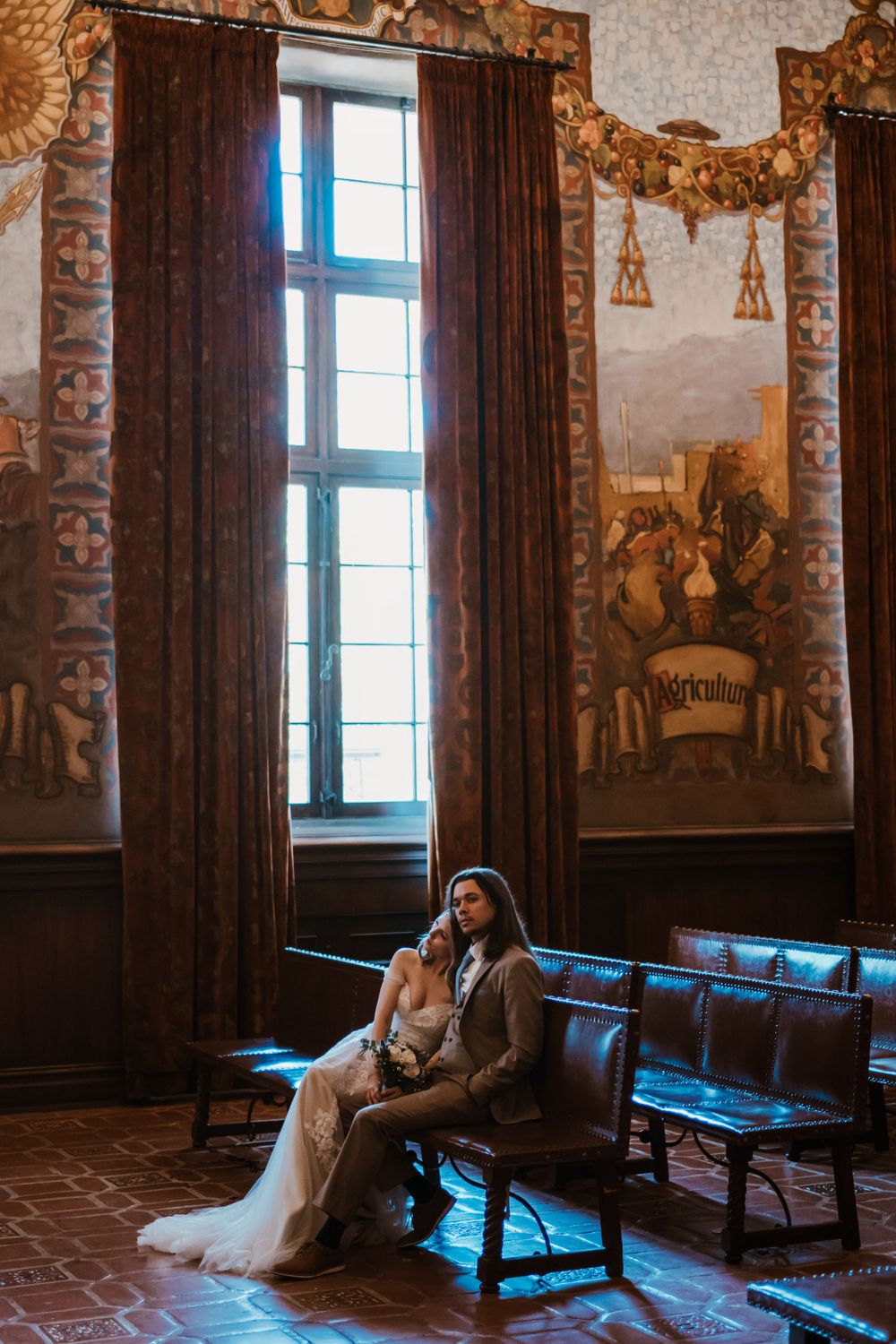 Two people sit together on an antique bench in an ornate room with tall windows and decorated walls featuring murals.