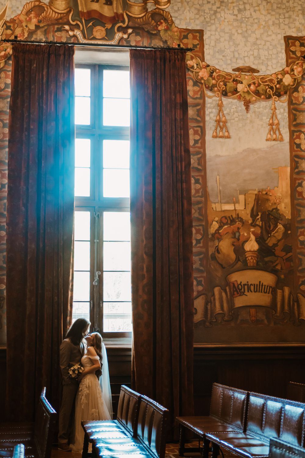 A couple stands by a tall window in an ornate historic room with dark curtains and decorative wall murals.