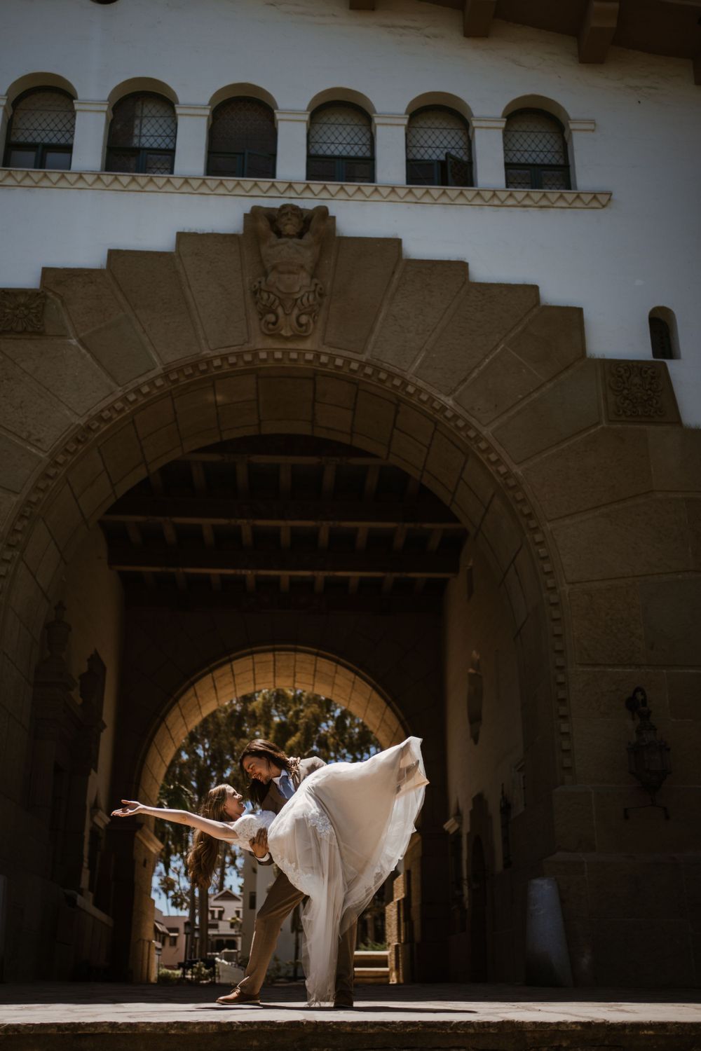 A dancer in a white dress performs a graceful arabesque pose under an ornate Spanish colonial stone archway.
