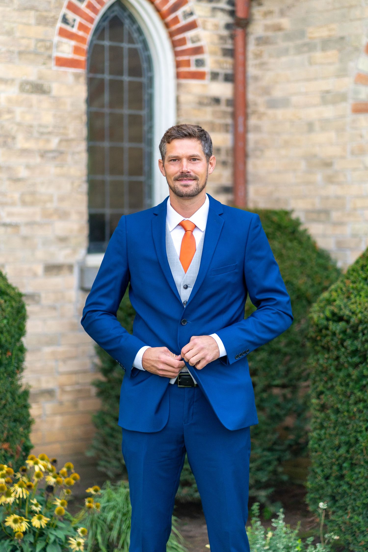 Groom in royal blue suit with orange tie stands in front of church window.