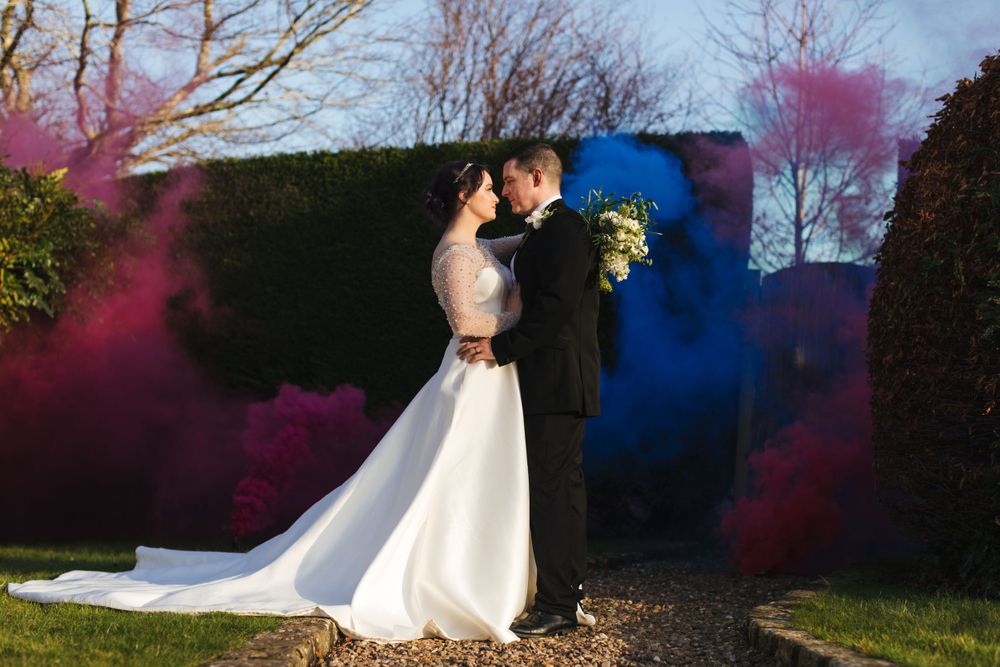 A tender wedding moment captured against dramatic pink and blue smoke effects in a garden setting.