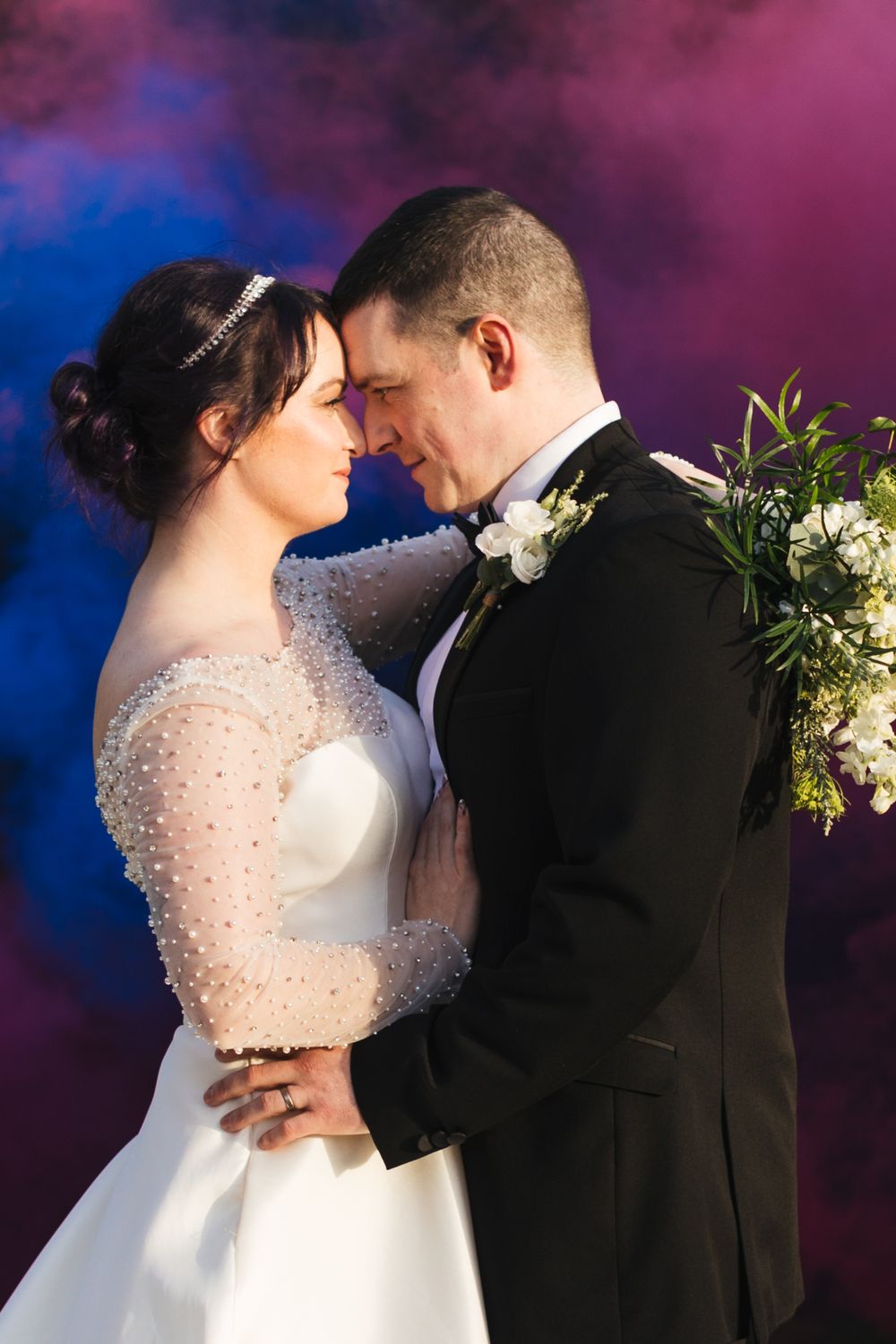 An intimate wedding portrait with pink and blue smoke effects featuring elegant formal attire.